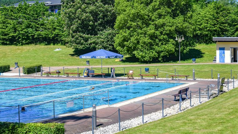 Ein Schwimmbad in einer grünen Umgebung mit Sonnenliegen und einem Sonnenschirm. Umgeben von Bäumen und einem klaren Himmel.