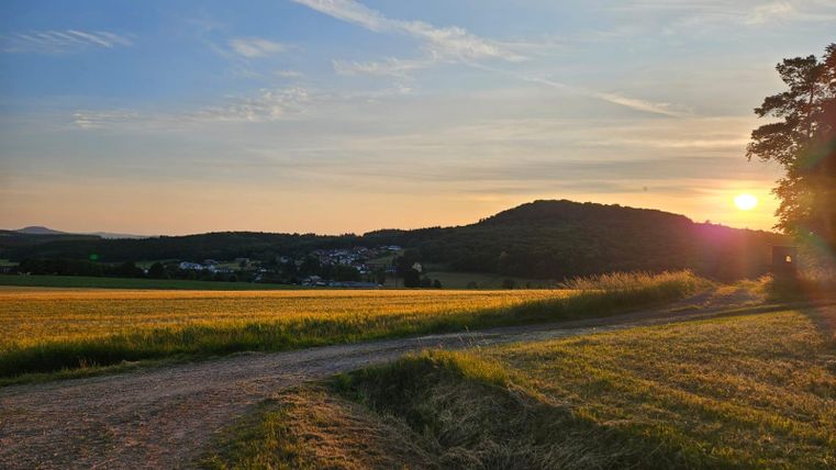 Eine malerische Landschaft mit sanften Hügeln und goldenen Feldern. Die Sonne geht langsam hinter den Bergen unter und färbt den Himmel in warmen Farbtönen.
