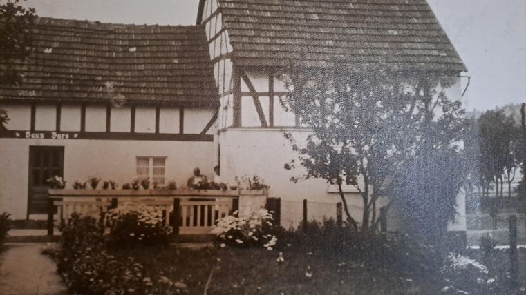 Ein traditionelles Fachwerkhaus mit einem Garten. Im Vordergrund sind Blumen und ein kleiner Weg zu sehen.