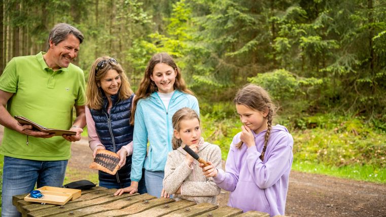 Eine Familie steht an einem Tisch im Wald und betrachtet gemeinsam Werkzeuge. Die Kinder scheinen interessiert und besprechen etwas miteinander.