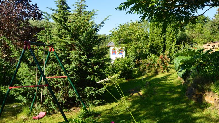 Ein schöner Garten mit dichten Bäumen und einer Schaukel. Im Hintergrund ist ein Haus sichtbar, umgeben von grüner Vegetation.