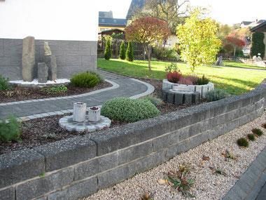 Ein schön angelegter Garten mit einer gepflasterten Auffahrt und verschiedenen Pflanzen. Auf der linken Seite stehen dekorative Steine und Betonringe.