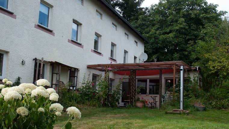 Ein gemütliches Haus mit einer überdachten Terrasse. Der Garten ist grün und blüht mit weißen Blumen.