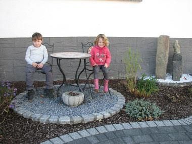 Zwei Kinder sitzen an einem kleinen Tisch in einem Garten. Um sie herum sind Pflanzen und Steine angeordnet.