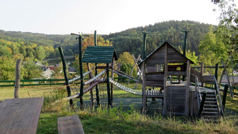 Ein Spielplatz mit Holzspielgeräten und Seilbrücken in einer hügeligen Landschaft. Umgeben von grünen Bäumen und sanften Hügeln.