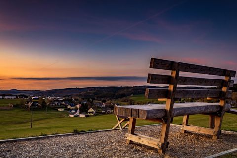 Eine Holzbank mit Blick auf eine malerische Landschaft bei Sonnenuntergang. Im Hintergrund sind Hügel und ein kleines Dorf zu sehen.