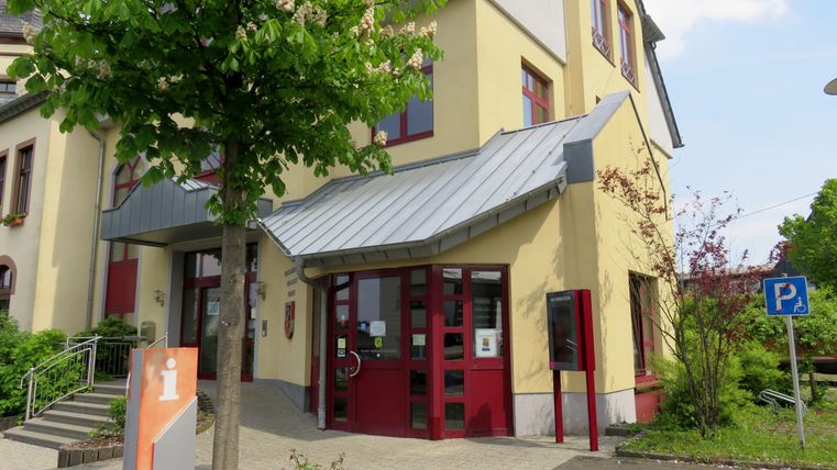 Ein modernes Gebäude mit roter Eingangstür und einem begrünten Außenbereich. Parkmöglichkeiten sind in der Nähe vorhanden.