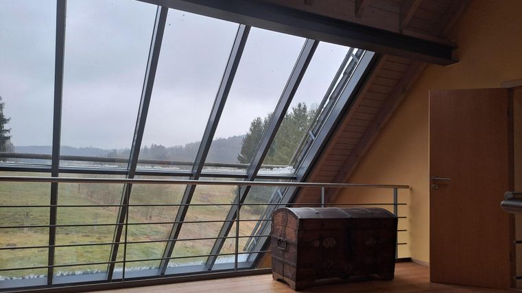Ein modernes Zimmer mit großen Fenstern und Blick auf eine grüne Landschaft. Im Raum steht eine rustikale Truhe aus Holz.
