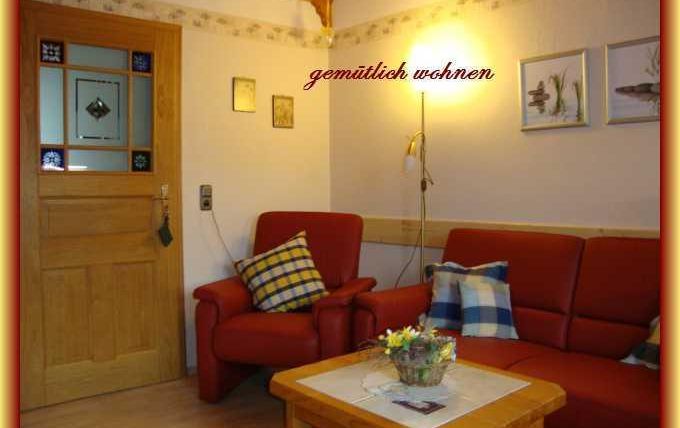 Ein gemütliches Wohnzimmer mit roten Sofas und einem Holztisch. An der Wand hängen Bilder, und eine Lampe sorgt für warmes Licht.