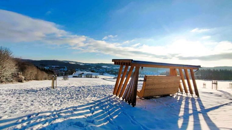 Eine Holzschaukel steht in einer verschneiten Landschaft. Im Hintergrund sind sanfte Hügel und ein klarer blauer Himmel zu sehen.