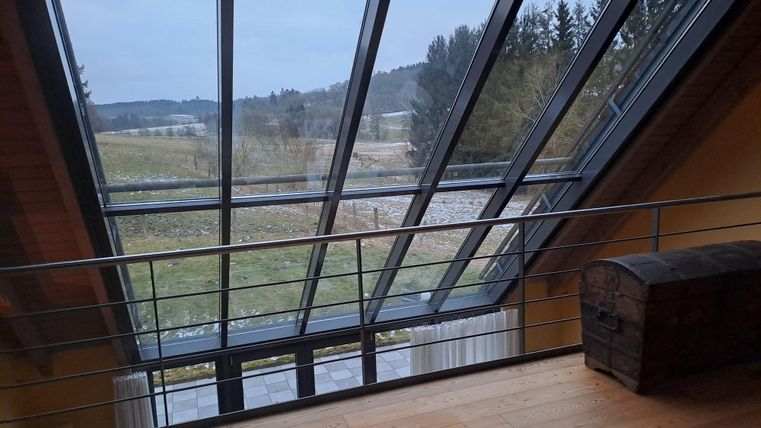 Ein moderner Raum mit großen Glasfenstern, die einen Blick auf die grüne Landschaft bieten. Der Boden ist aus hellem Holz, und in der Ecke steht eine alte Truhe.