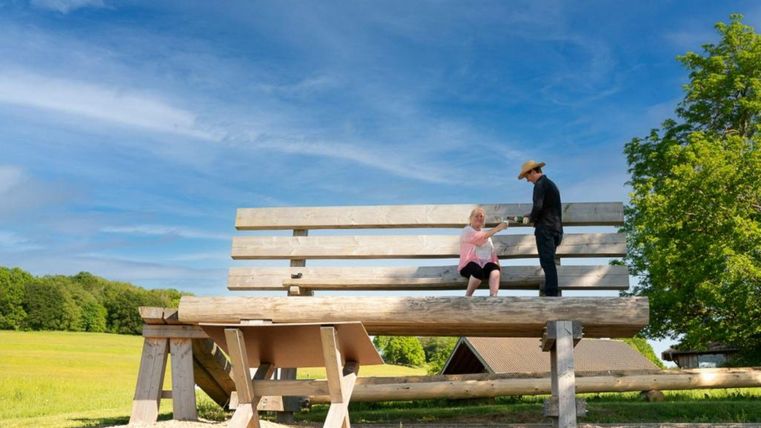 Eine große Holzbank steht in einer grünen Landschaft unter einem blauen Himmel. Zwei Figuren sind auf der Bank; eine sitzt und die andere steht daneben.