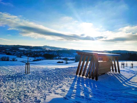 Eine Winterlandschaft mit schneebedeckten Feldern und einem Holzunterstand. Die Sonne scheint über den Hügeln und wirft lange Schatten.