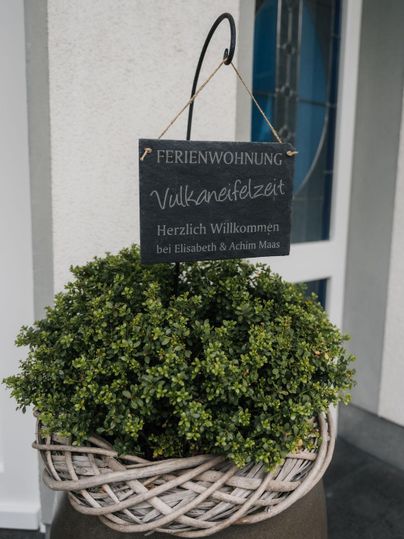 Ein Willkommensschild für eine Ferienwohnung namens "Vulkaniefelzeit" hängt an einer Pflanze in einem Körbchen. Die Atmosphäre ist gemütlich und einladend.