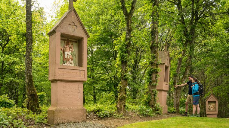 Ein Wanderer steht in einem Waldweg und betrachtet kunstvolle Nischen mit Figuren. Umgeben von grünen Bäumen und einer ruhigen Umgebung vermittelt die Szene eine friedliche Stimmung.