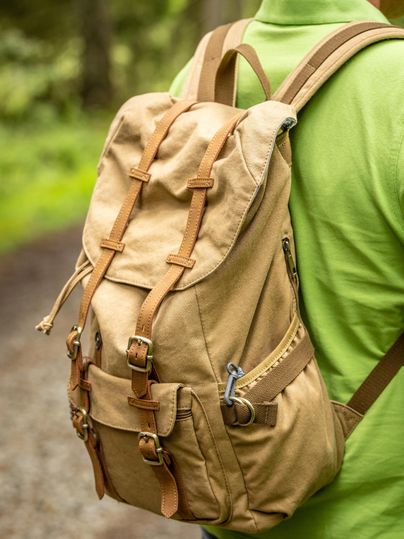 Ein beige Rucksack mit braunen Lederriemen, getragen von einer Person in einem grünen T-Shirt. Die Szene zeigt einen Waldweg im Hintergrund.