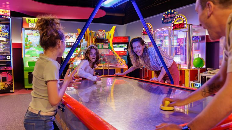 Eine Gruppe von Freunden spielt Air-Hockey in einer bunten Spielhalle. Es gibt viele Spielautomaten im Hintergrund.