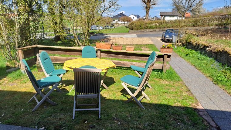 Ein gemütlicher Garten mit einem runden Tisch und sechs Stühlen. Im Hintergrund sind Wohnhäuser und ein auto sichtbar.