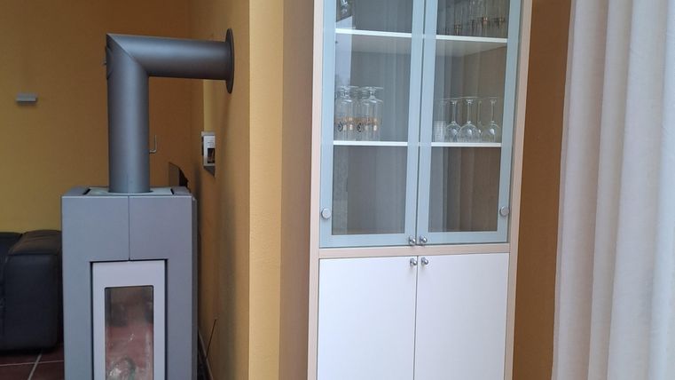 Ein moderner Glaskasten steht an einer wandfarbenen Wand. Neben ihm befindet sich ein Kamin mit Holzkorb.