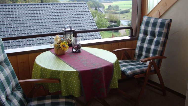 Ein gemütlicher Balkon mit einem runden Tisch und zwei Stühlen. Auf dem Tisch steht eine Blumenvase und eine kleine Dekoration, während die Aussicht ins Grüne einladend wirkt.