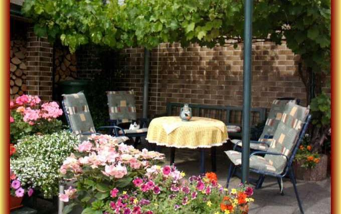 Ein gemütlicher Gartenbereich mit einem runden Tisch und Stühlen, umgeben von bunten Blumen. Der Bereich ist von grünen Pflanzen beschattet und lädt zum Entspannen ein.