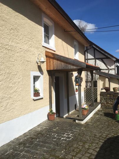 Ein charmantes Haus in hellen Farben mit einem Holzvordach. Es gibt Blumen in Töpfen und einen klaren blauen Himmel im Hintergrund.