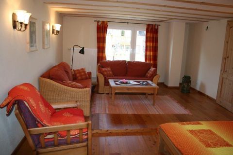 Ein gemütliches Wohnzimmer mit rot-orangenen Sofas und einem Tisch in der Mitte. Große Fenster bringen viel Licht in den Raum.