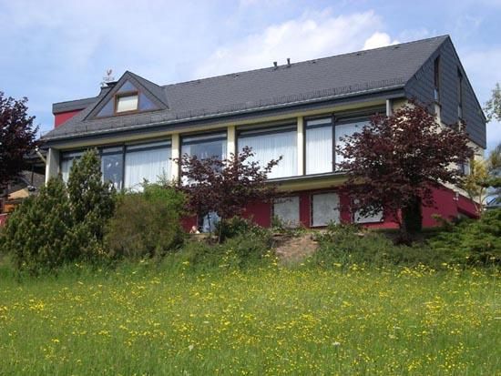 Ein modernes Haus mit großen Fenstern und einem schrägen Dach. Umgeben von Grünflächen und blühenden Bäumen.