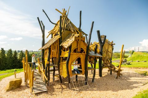 Ein Spielplatz mit einem großen Holzturm und vielen Kletterelementen. Kinder spielen und erkunden die Struktur in einer grünen Umgebung.