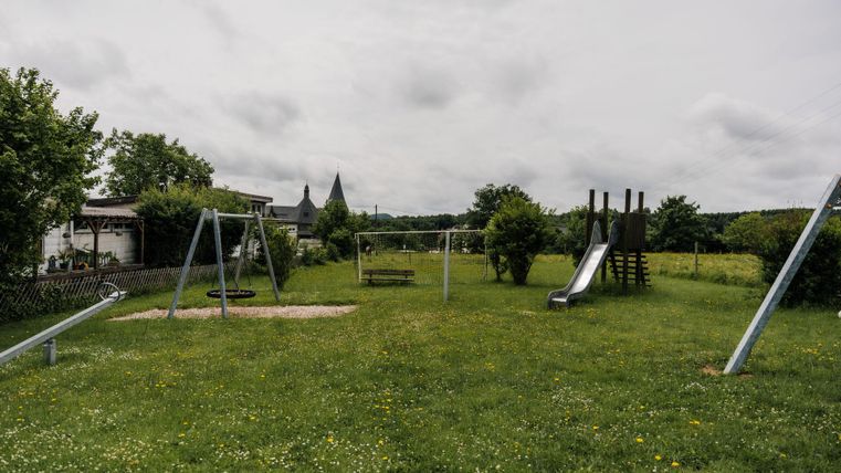 Ein Spielplatz mit Schaukeln, einer Rutsche und einer Kletterstruktur. Die Wiese ist grün und es gibt einige Bäume im Hintergrund.