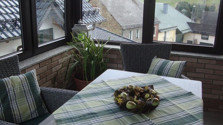 Ein gemütlicher Wintergarten mit einem Tisch und zwei Stühlen. Auf dem Tisch liegt ein Dekorationsteller mit Kräutern und einer Tischdecke im Karomuster.