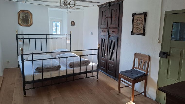 Ein helles Schlafzimmer mit einem Metallbett, einem Holzstuhl und einem großen Schrank. Die Wände sind weiß und der Boden ist aus Holz.