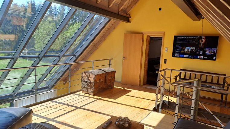 Ein modernes Zimmer mit großen Fenstern und Holzbalken. Es gibt ein Fernseher, eine Couch und einen Tisch aus Holz.
