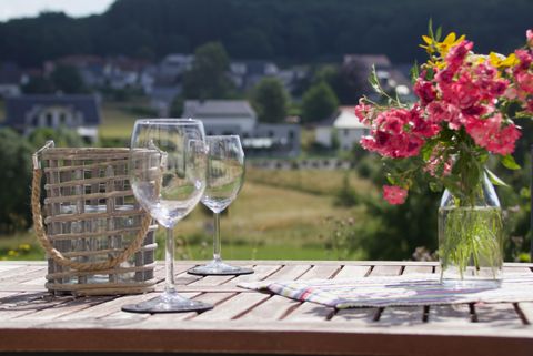 Ein Tisch im Freien mit zwei Weingläsern und einem Korb. Neben den Gläsern steht eine Vase mit bunten Blumen und im Hintergrund sind grüne Felder und Häuser zu sehen.