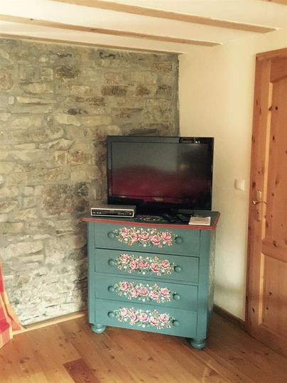 Ein gemütliches Zimmer mit einer Steinmauer und einem hübschen, bemalten Schrank. Darauf steht ein Fernseher und es gibt einen warmen Holzfußboden.