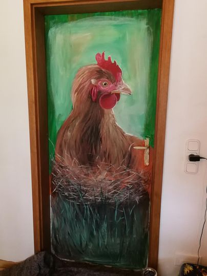 Eine bemalte Tür zeigt ein Huhn, das in einem Nest sitzt. Der Hintergrund ist grün und vermittelt eine ländliche Atmosphäre.