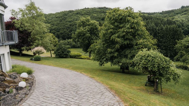 Ein gepflegter Garten mit üppigem Grün und mehreren Bäumen. Der Weg führt sanft durch die Landschaft, umgeben von Hügeln.