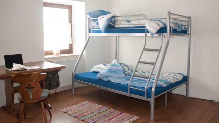 Ein einfaches Zimmer mit einem Etagenbett und einem Schreibtisch. Die Wände sind hell und der Boden ist aus Holz.