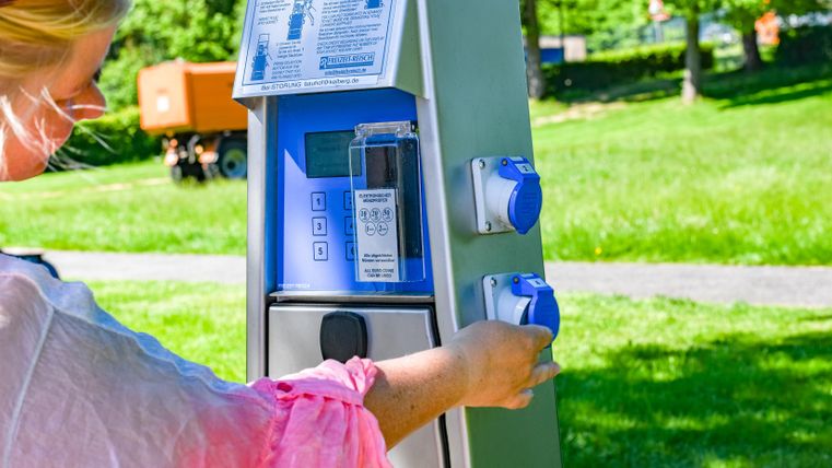 Eine Person bedient einen Parkautomaten in einem grünen Park. Der Automat hat blaue Knöpfe und eine informative Anzeige.