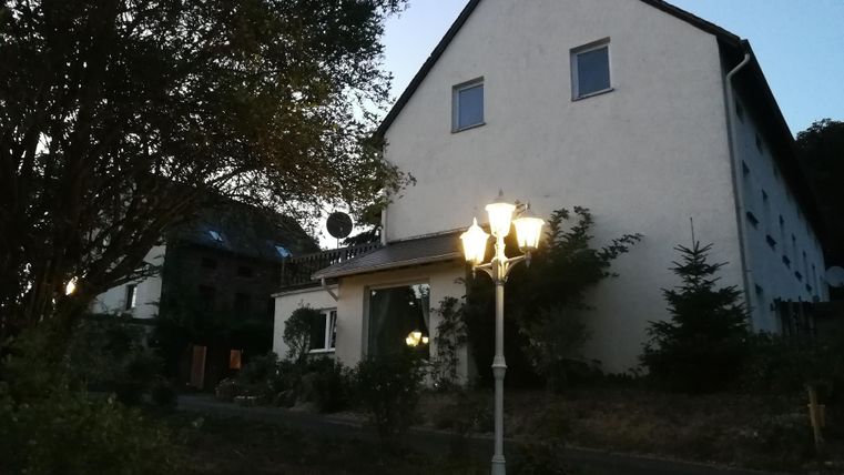 Ein gemütliches Haus in der Dämmerung, umgeben von Bäumen und Sträuchern. Eine Straßenlampe beleuchtet den Eingang.