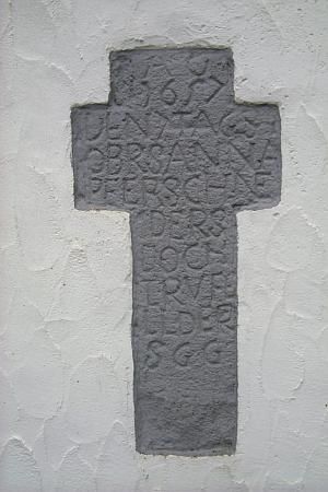 Ein graues Kreuz aus Stein mit einer gravierten Inschrift. Das Kreuz ist an einer weißen Wand angebracht.