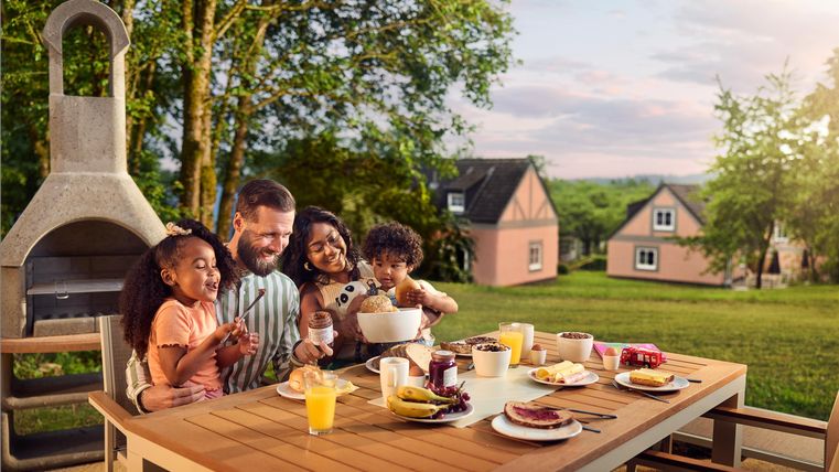 Eine glückliche Familie sitzt am Tisch im Freien und genießt ein Frühstück. Im Hintergrund sind Bäume und gemütliche Häuser zu sehen.