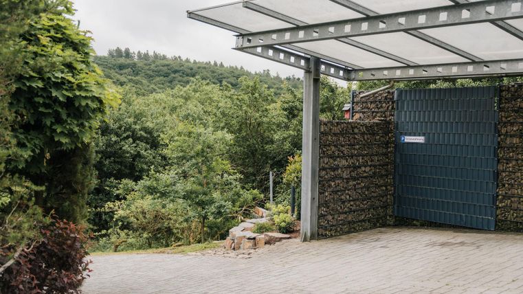 Ein moderner Carport mit Glasdach steht vor einem Farbton. Im Hintergrund sind grüne Bäume und Hügel zu sehen.