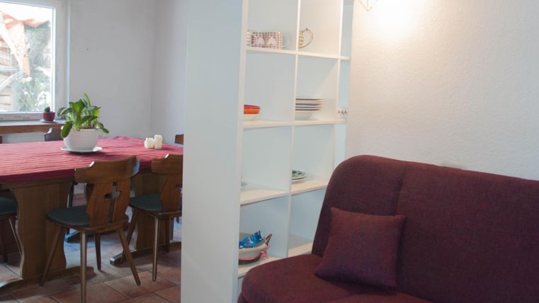 Ein gemütliches Wohnzimmer mit einem lila Sofa und einem Bücherregal. Im Hintergrund ist ein Esstisch mit Stühlen und einer Pflanze zu sehen.
