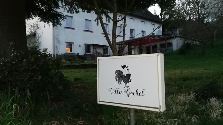 Ein Schild mit der Aufschrift "Villa Gechel" steht vor einem großen, weißen Haus. Im Hintergrund sind Bäume und ein gemütlicher Garten sichtbar.