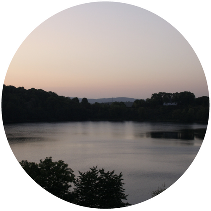 Ein ruhiger See mit sanften Wellen und einer sanften Abenddämmerung. Umgeben von Bäumen und sanften Hügeln im Hintergrund.