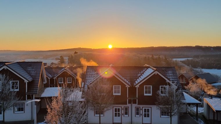 Ein malerischer Sonnenaufgang über verschneiten Häusern. Die Szene zeigt eine ruhige Winterlandschaft mit sanften Hügeln im Hintergrund.