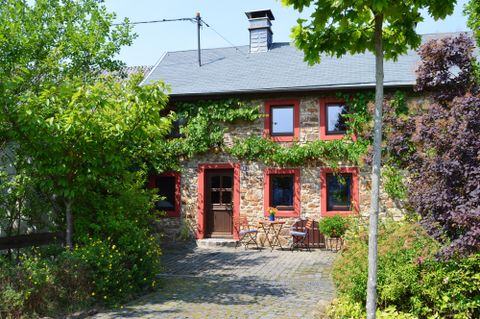 Ein charmantes Steinhaus mit roten Fenstern und Tür, umgeben von Pflanzen. Der Außenbereich ist von grünem Laub und blühenden Sträuchern geprägt.
