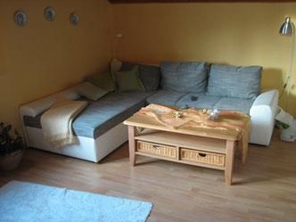Ein gemütliches Wohnzimmer mit einer hellen Couch und einem Holztisch. Auf dem Tisch befindet sich eine dekorative Tischdecke und einige kleine Deko-Elemente.