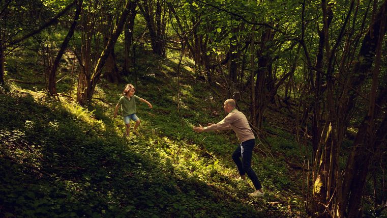 Ein Vater spielt mit seinem Kind in einem grünen Wald. Sie bewegen sich fröhlich durch das Unterholz.